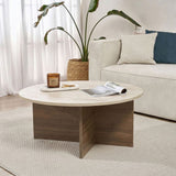 Ronde salontafel met travertin blad en donker houten onderstel in kruisvorm. Op de achtergrond bank en plant.